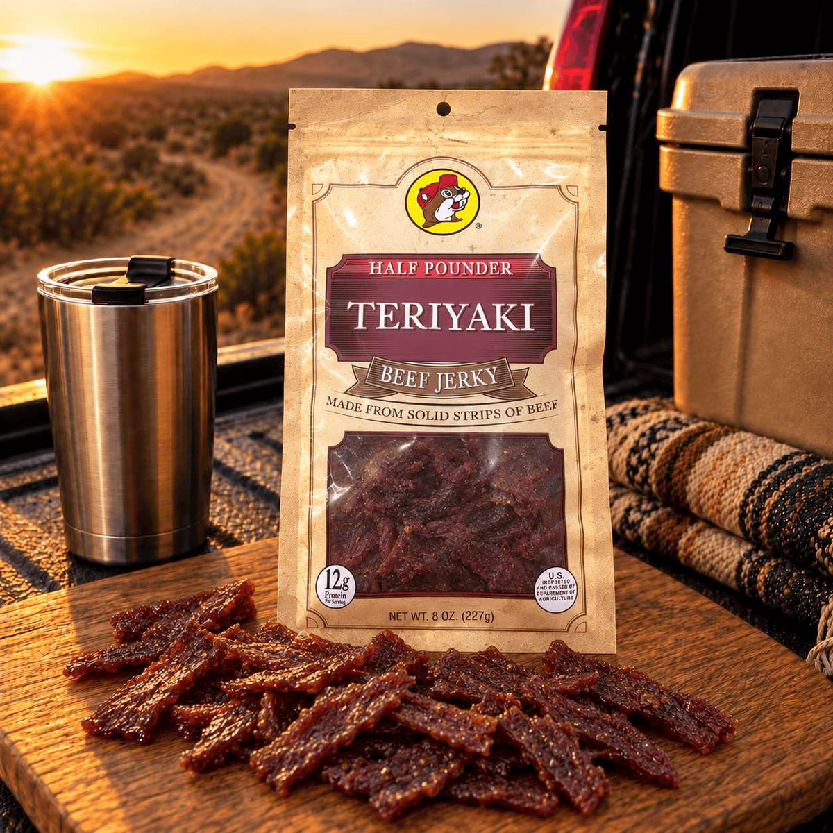 A bag of Teriyaki Beef Jerky is displayed on a wooden cutting board outdoors at sunset, with sliced jerky pieces scattered in front. A stainless steel tumbler, a cooler, and a striped blanket are visible in the background, with a desert landscape and vehicle tailgate behind them.